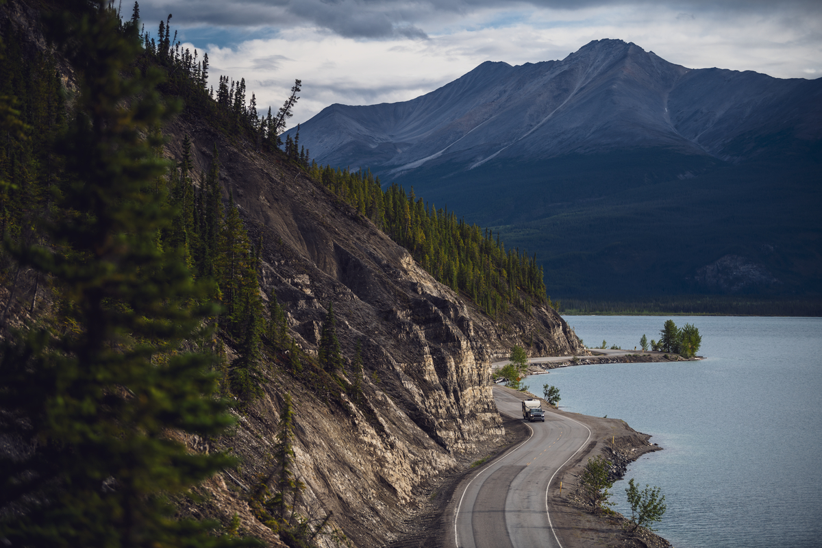 A two lane paved road winds between a forested hillside and a lake with mountains in the background on the drive to alaska