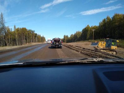 a construction zone on a highway in alaska. The road is graded gravel and there is a small bulldozer next to the road. Trucks and cars are goign through the construction.