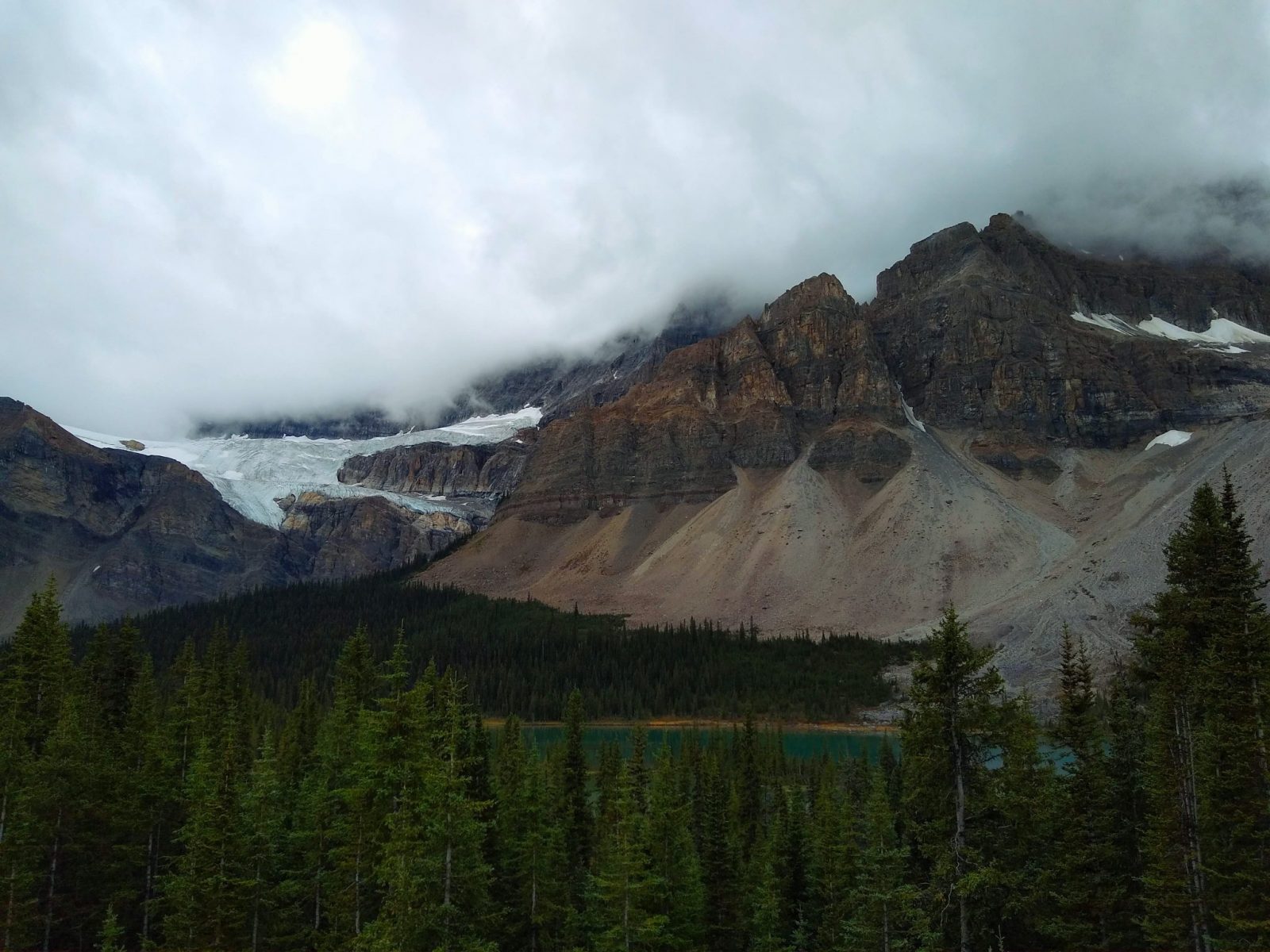 mountains with a hanging glacier above a lake and forests on a dark and overcast day
