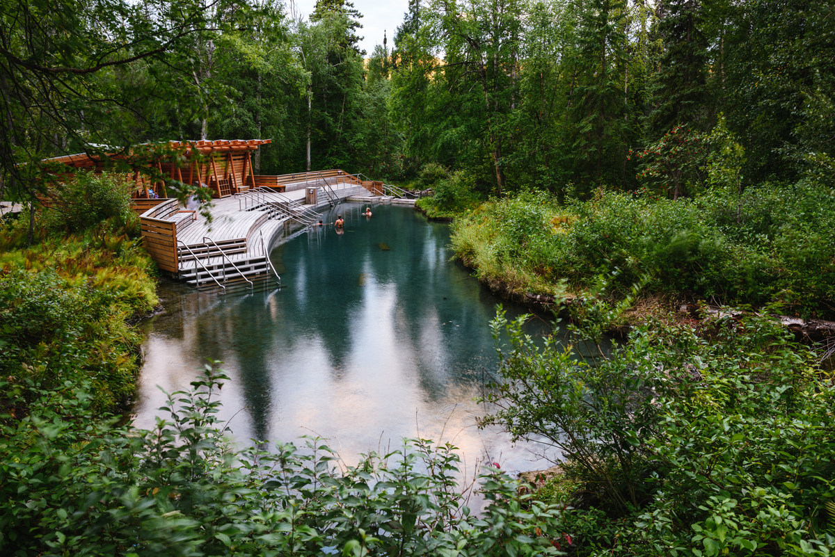 Along the drive to Alaska, Liard Hot Springs is a hot pool with platforms and stairs to help visitors get in the water. There is a wooden building for changing next to the pool. The pool is surrounded by lush green forest and bushes