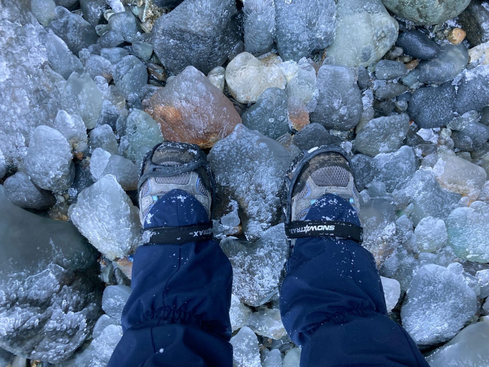 A person's lower legs standing on icy rocks. They are wearing hiking shoes and gaiters and microspikes