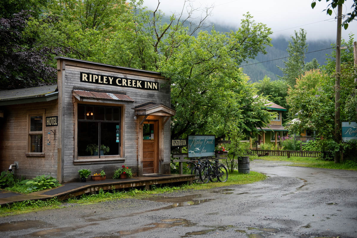 A small historic wooden building says Ripley Creek Inn. It's next to a gravel road in the forest near a small town on an overcast day