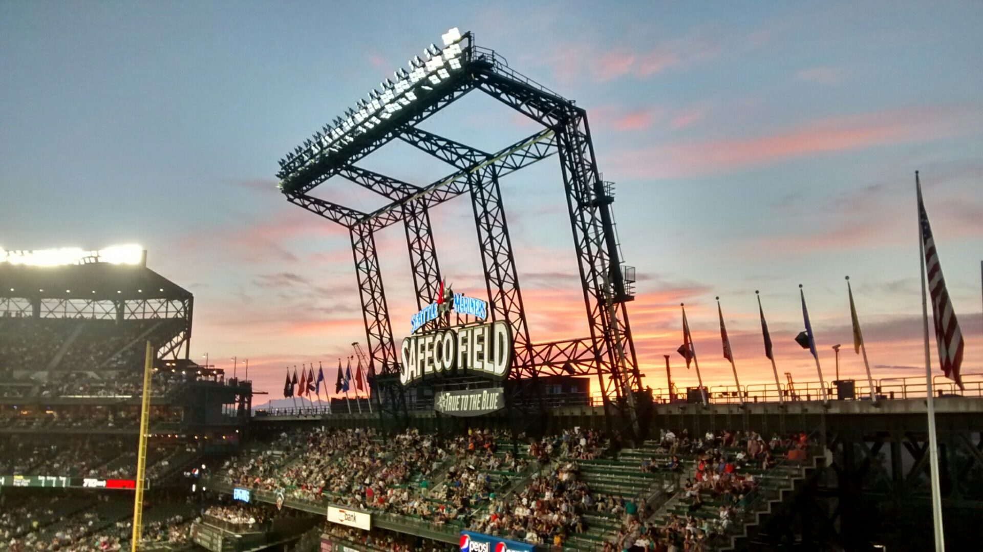 Safeco Field in Seattle. The sun is setting and the roof is open. The lights have just come on. There are fans and flags and a steel structure holding the lights.