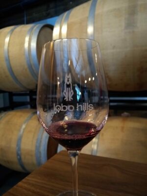 A wine glass with a bit of red wine in it. The wine glass has a logo on it and is on a wooden table in front of wine barrels