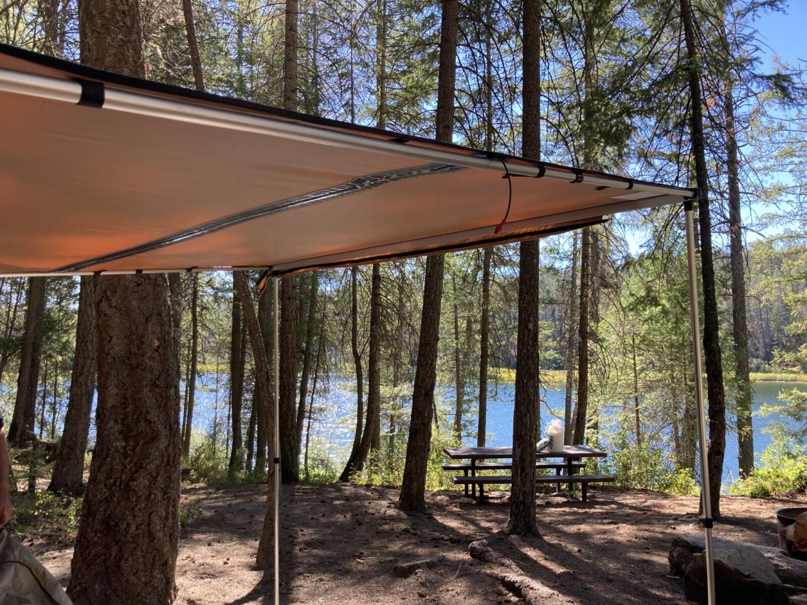 A roof rack awning set up and providing shade in a forested campground next to a lake