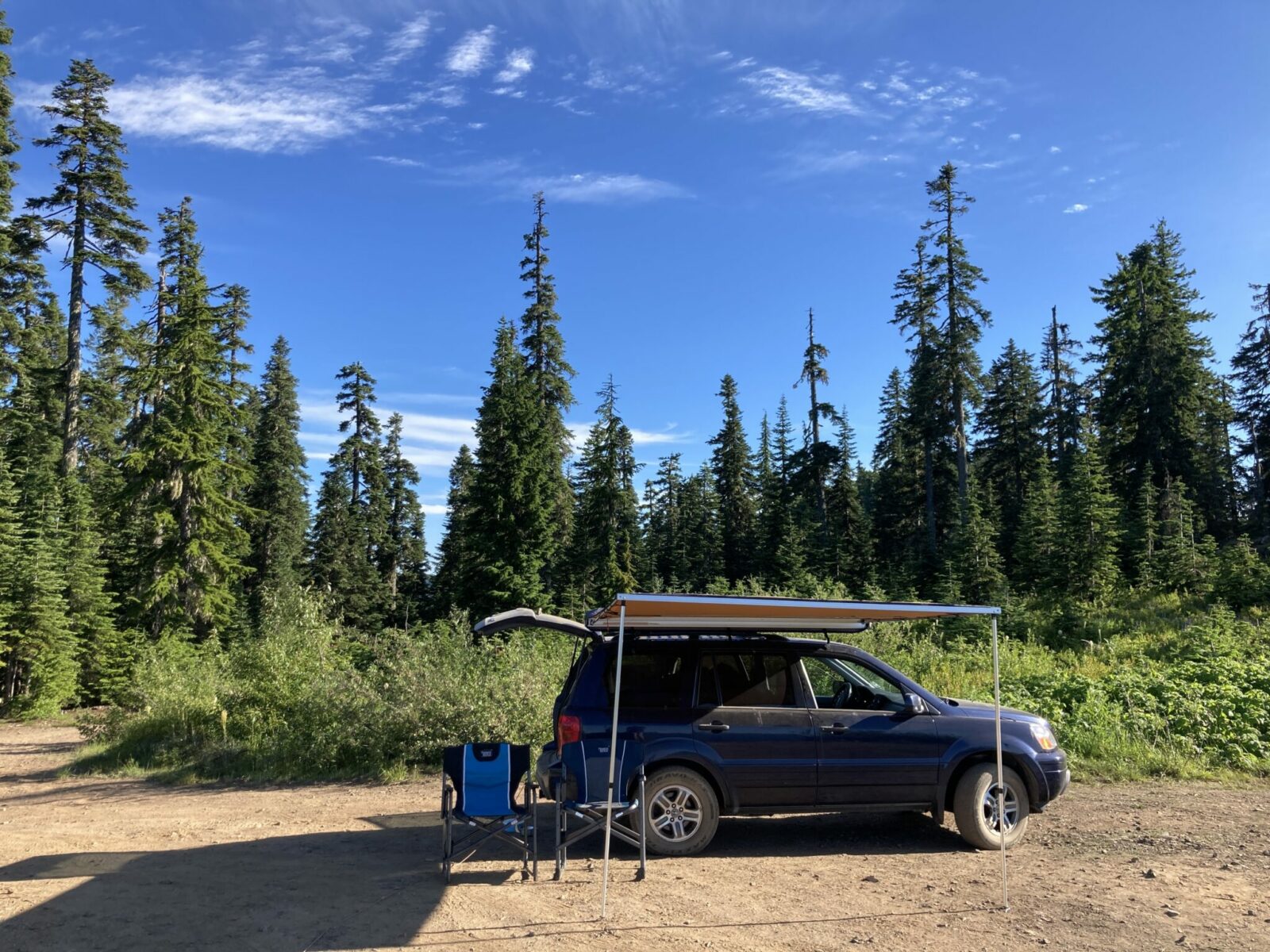 A blue SUV parked on a dirt open area in the forest surrounded by bushes. The rear hatch of the SUV is open and a roof top awning is set up on the side. There are two camp chairs sitting next to it.