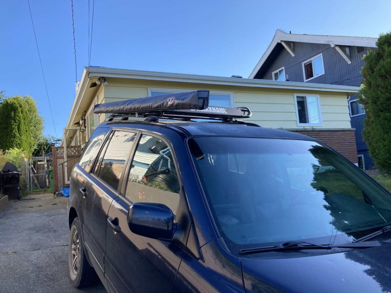 A blue SUV in a driveway in front of a yellow house. It has a roof rack and a roof rack awning rolled up on the top