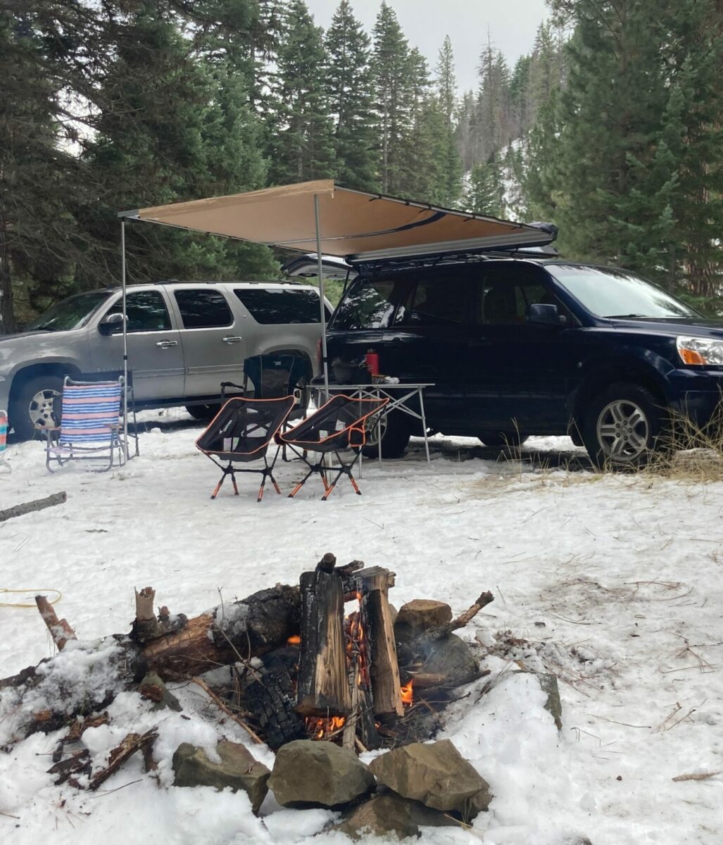 A gray SUV parked near a blue SUV. The blue SUV has a roof top awning set up on the side with a table and camp chairs under it. There is snow on the ground and in the foreground is a campfire