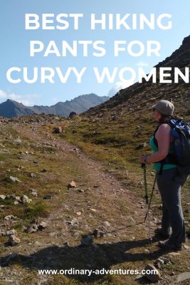 A trail in a meadow next to a higher mountain with mountains in the distance and a person on the trail wearing gray hiking pants, a green tank top, a hat and a backpack and carrying hiking poles. Text reads: best hiking pants for curvy women