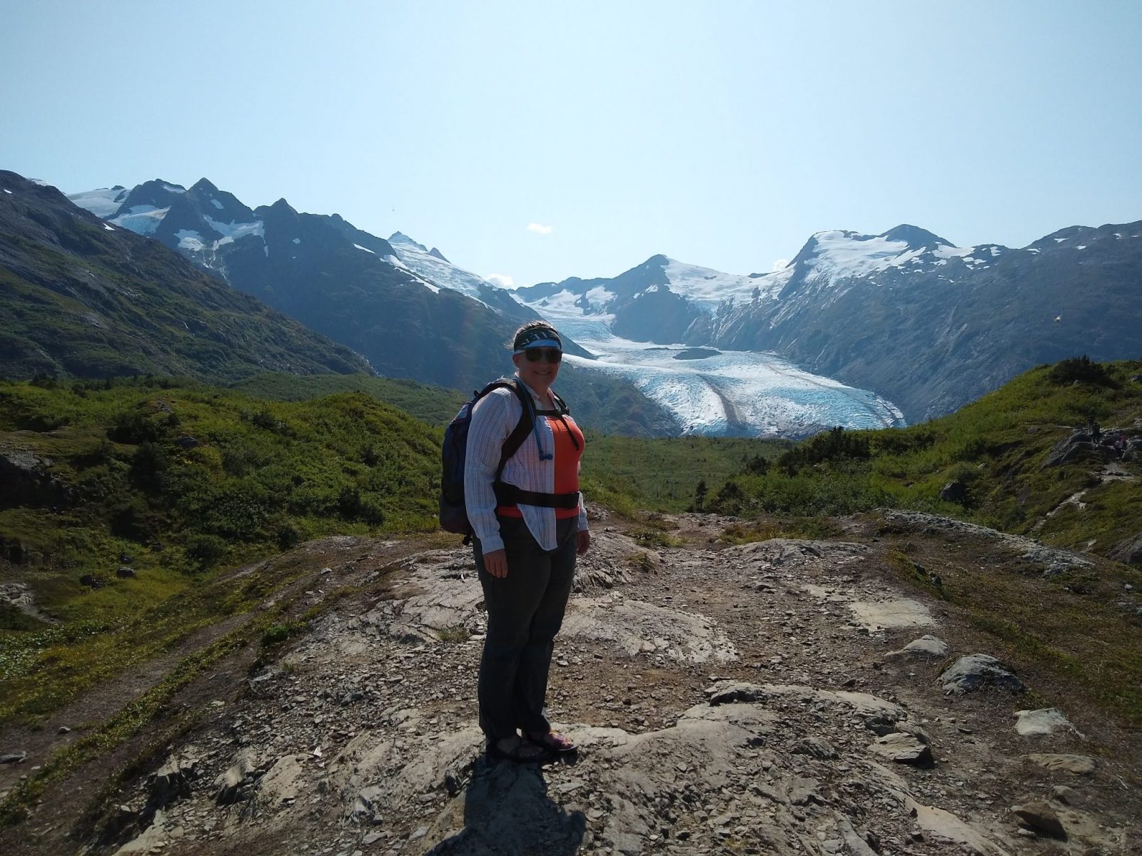 A woman on a rocky trail in a meadow with mountains and a glacier in the background wearing hiking pants, a tank top, a long sleeve shirt, a backpack and sunglasses