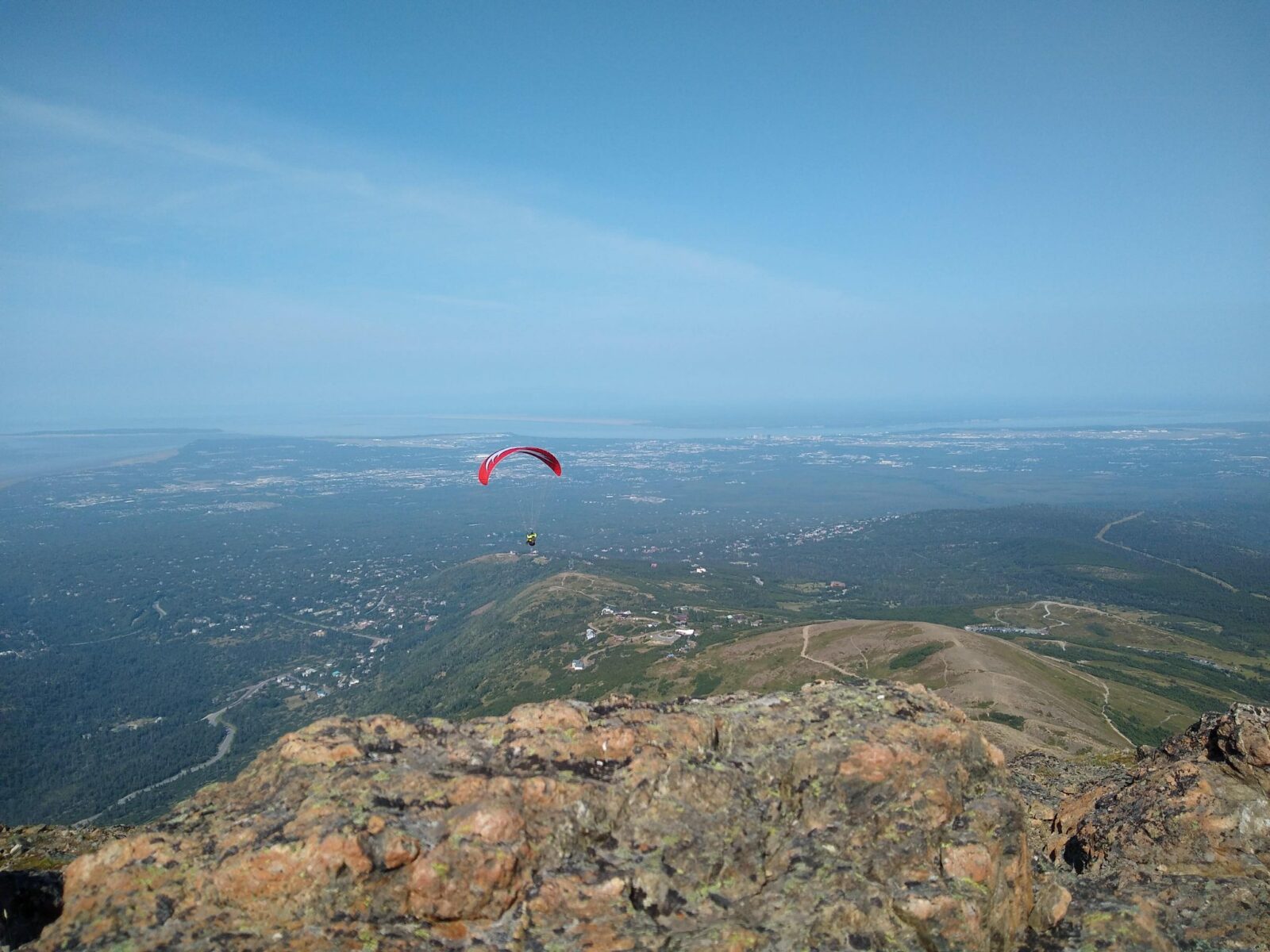 the rocky summit of flattop mountain with a trail below and houses and trees and roads far below. In the foreground is a paraglider with a red parachute