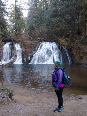 A woman at a waterfall on a cold day wearing hiking shoes, hiking pants, a purple coat, a backpack, gloves and a warm hat