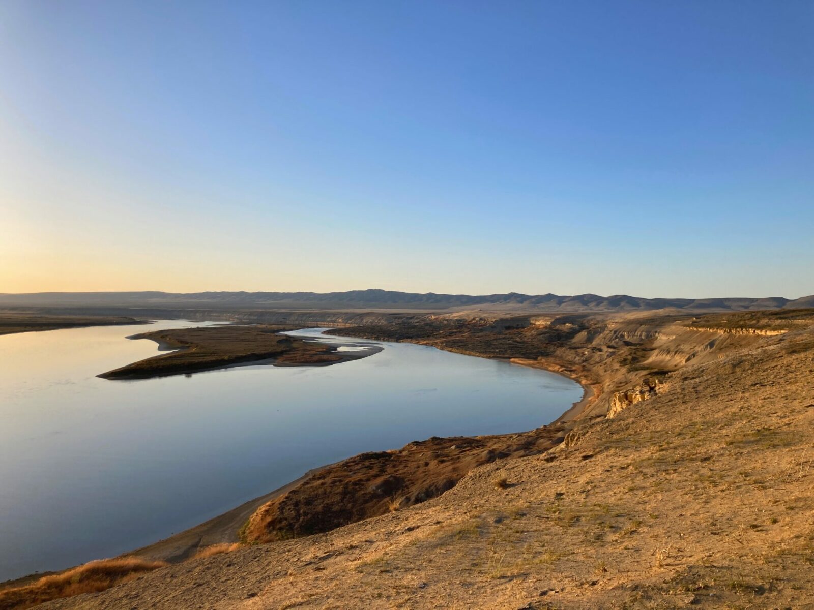 Hanford Reach is a section of the Columbia River in Eastern Washington. The river and an island in it are seen from the bluffs above, surrounded by bluffs and distant mountains at sunset on a sunny day