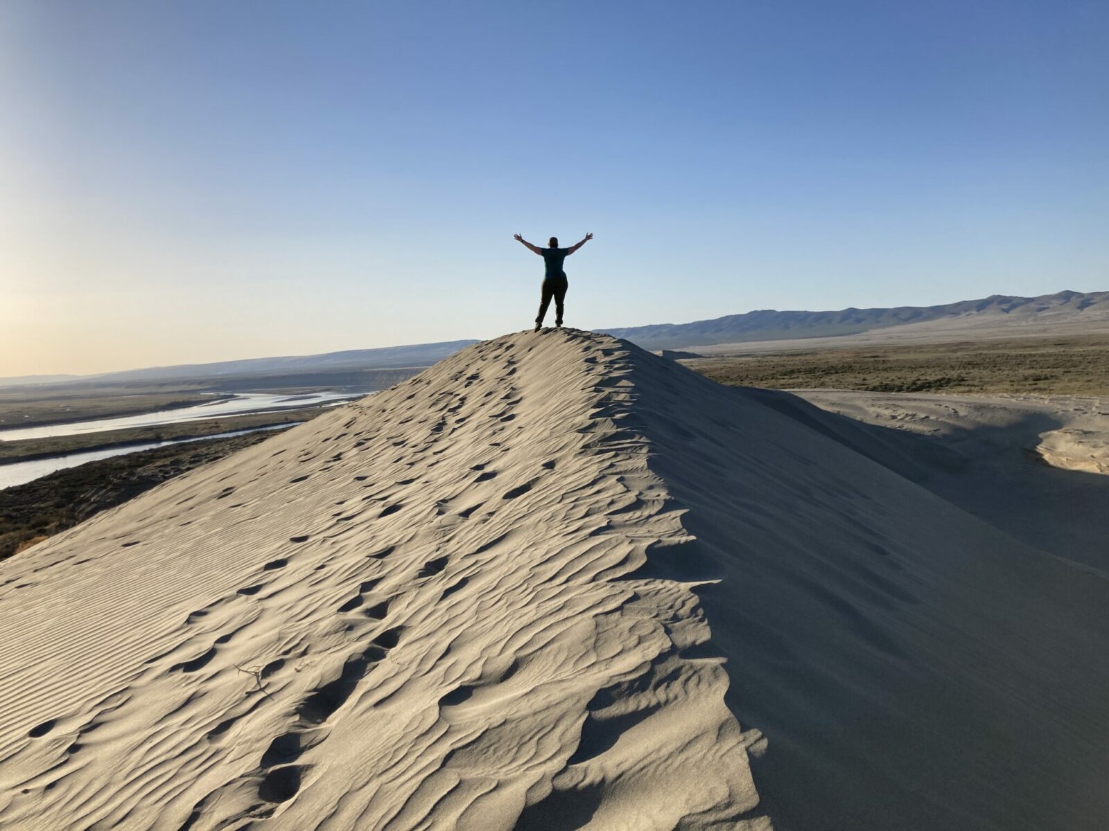 The top of a sand dune at Hanford Reach in a desert in late afternoon. A person stands on the top of the dune with their hands in the air. In the distance are brown mountains and a river