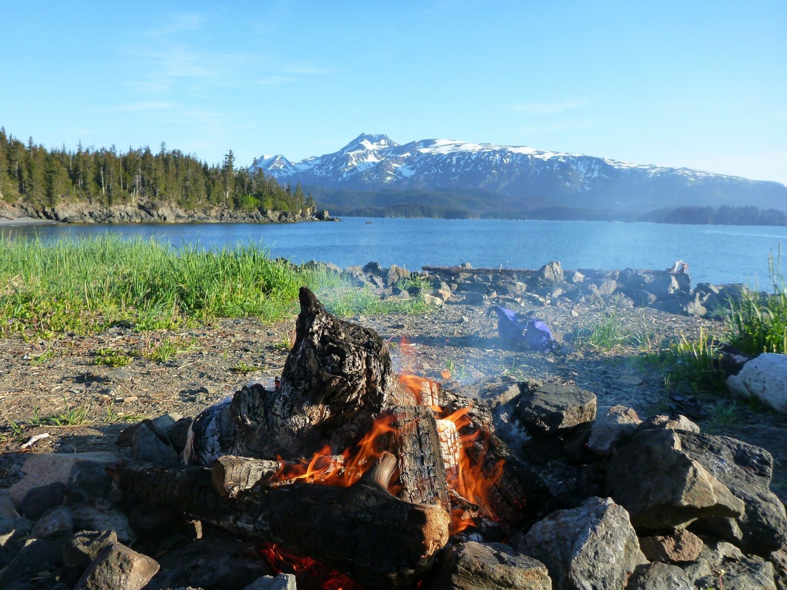 A campfire in the foreground on a rocky and grassy beach along a small bay. Across the bay there are forested hillsides and higher mountains with lingering snow