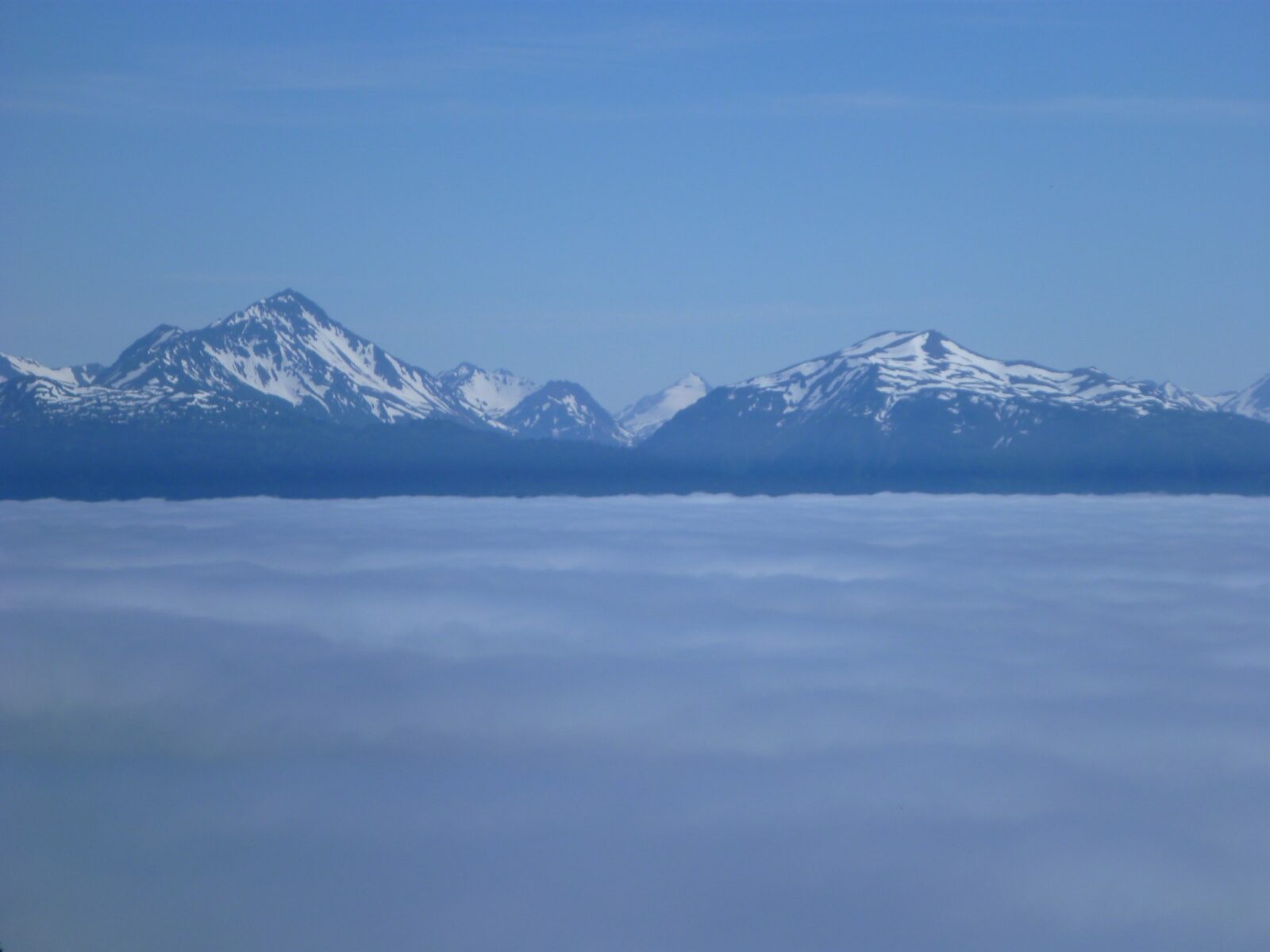 Mountains with lingering snow rise above a thick cloud layer on a sunny day