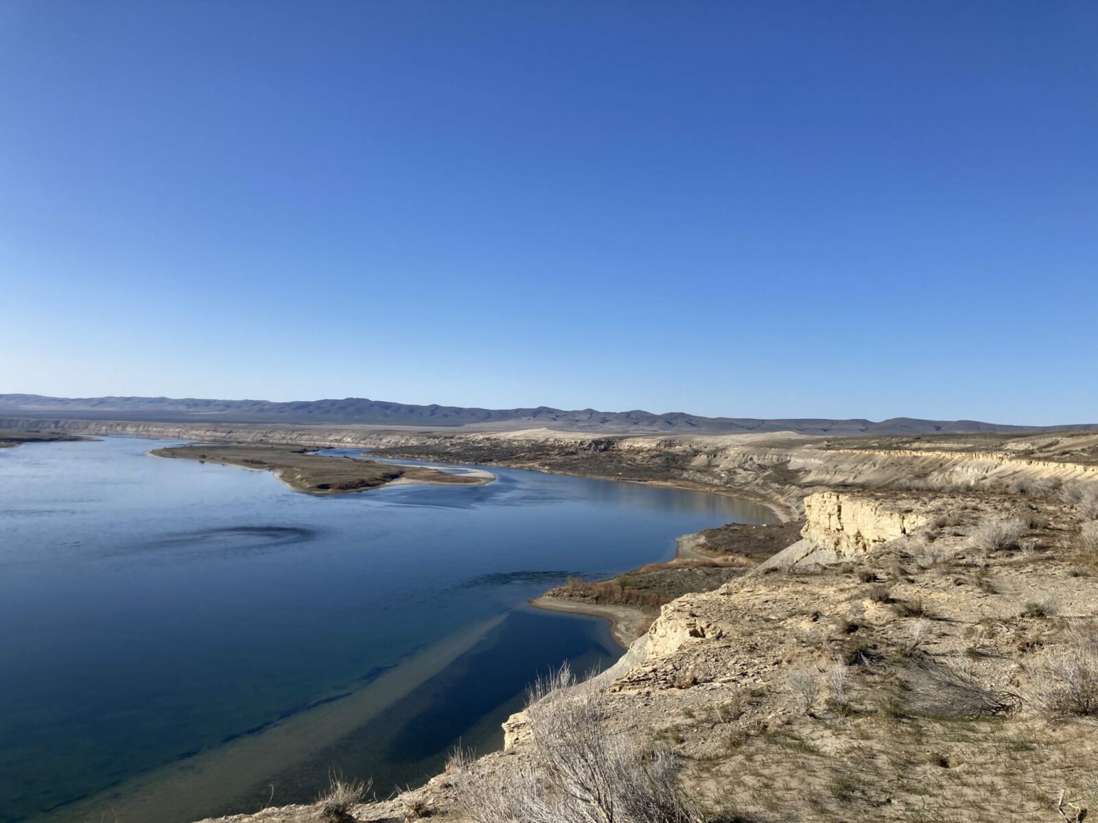 Hanford Reach is a section of the Columbia River in Eastern Washington. The river and an island in it are seen from the bluffs above, surrounded by bluffs and distant mountains on a sunny day