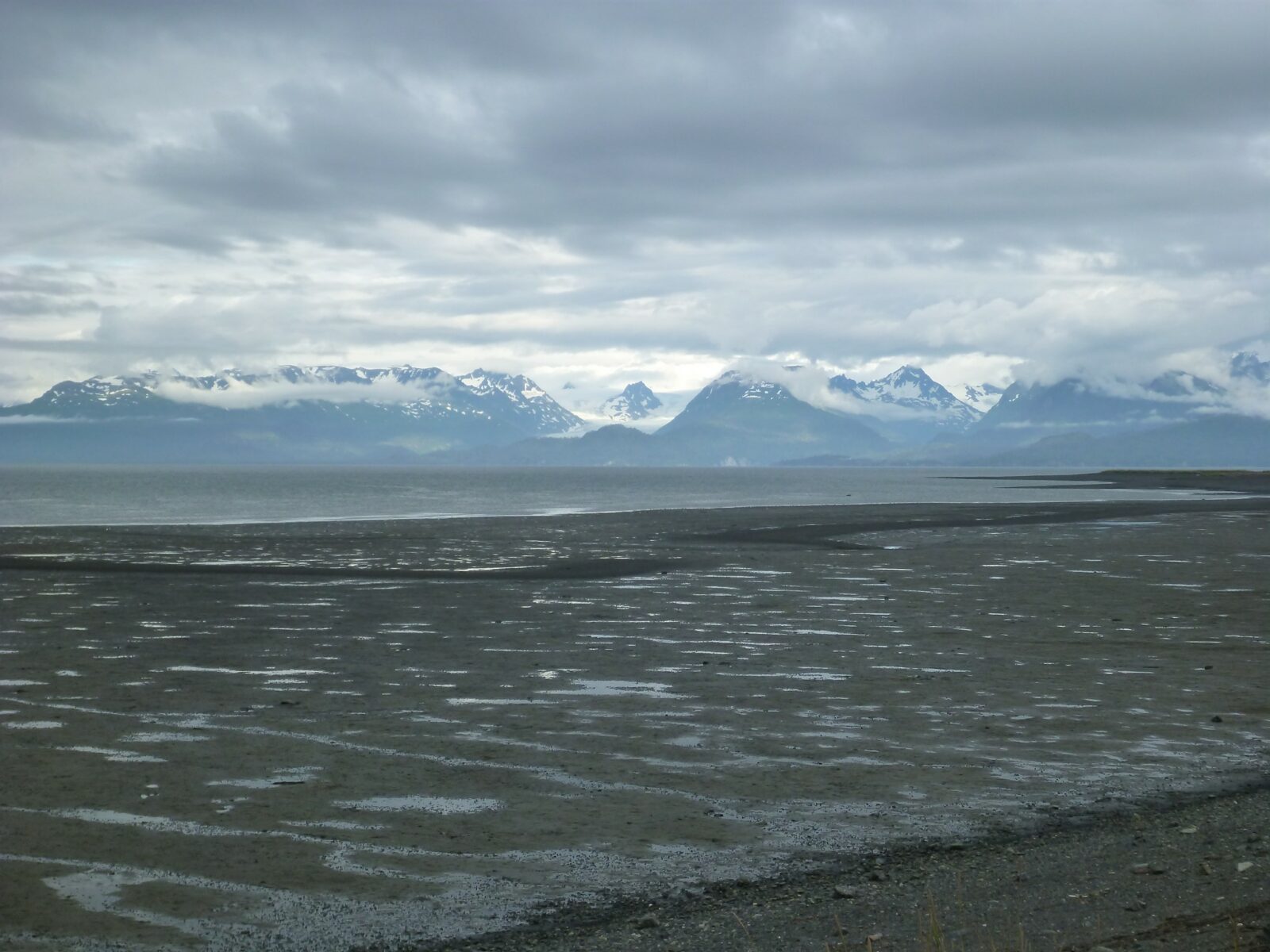 Low tide on the beach at gravelly Homer Spit. In the distance is gray water and mountains with snow and fog surrounding them on an overcast day