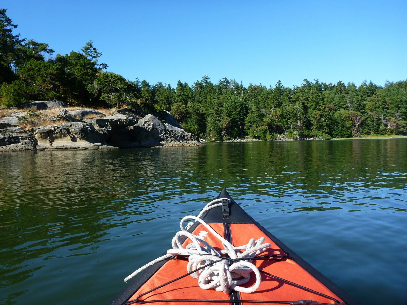 The front of a red kayak in Fossil Bay on Sucia Island. It's a sunny day and the forested island also has rocky outcroppings in the water