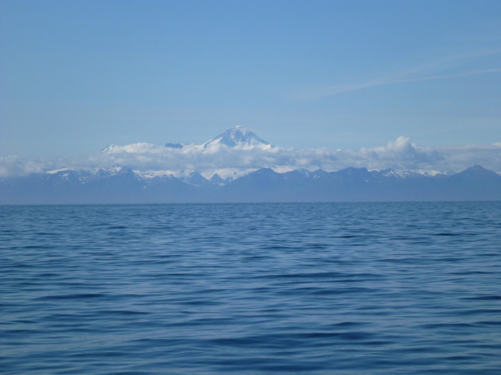 A high snow covered volcano surrounded by a few clouds with water in the foreground seen from the highway to Homer Alaska